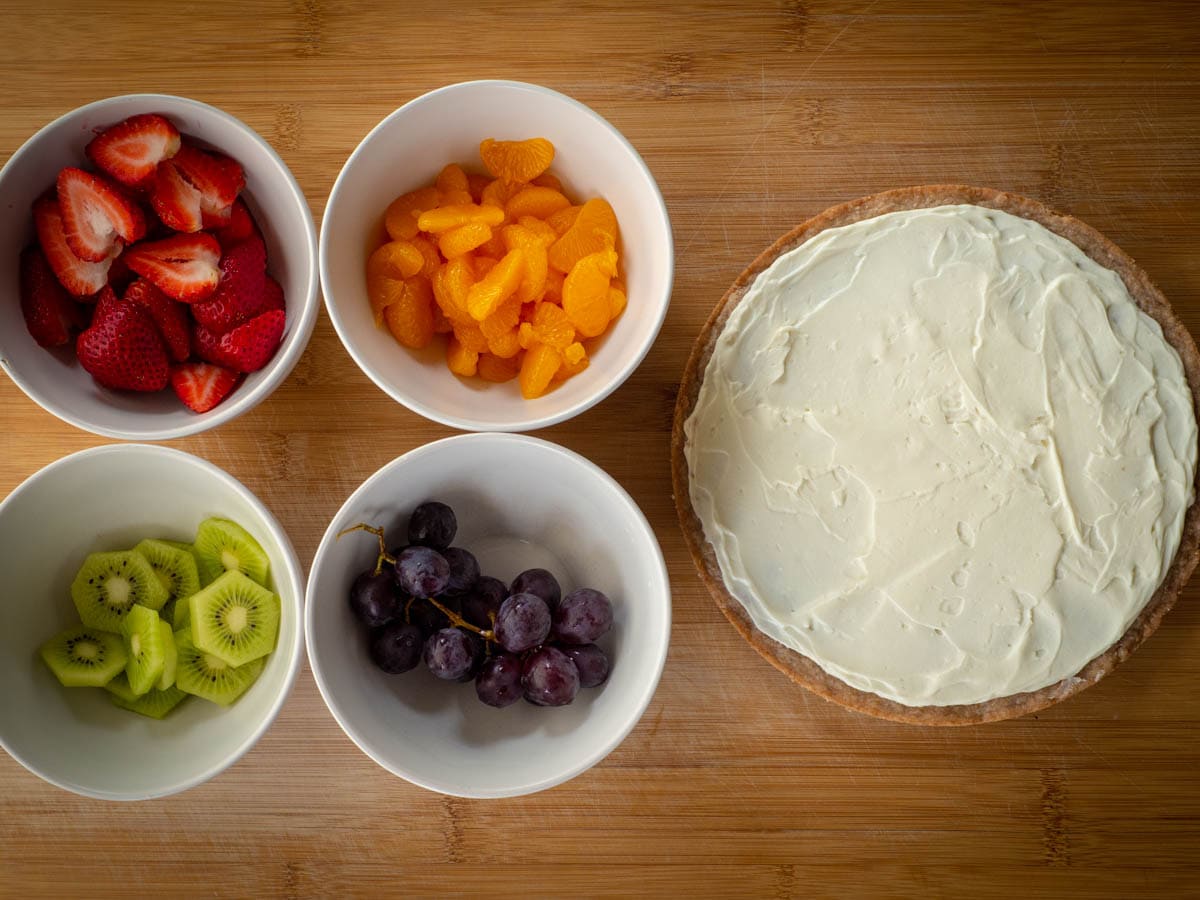 Bowl of fruit prepared for fruit pizza including sliced strawberries, oranges, kiwi and grapes. Crust topped with cream cheese sauce next to the bowls.