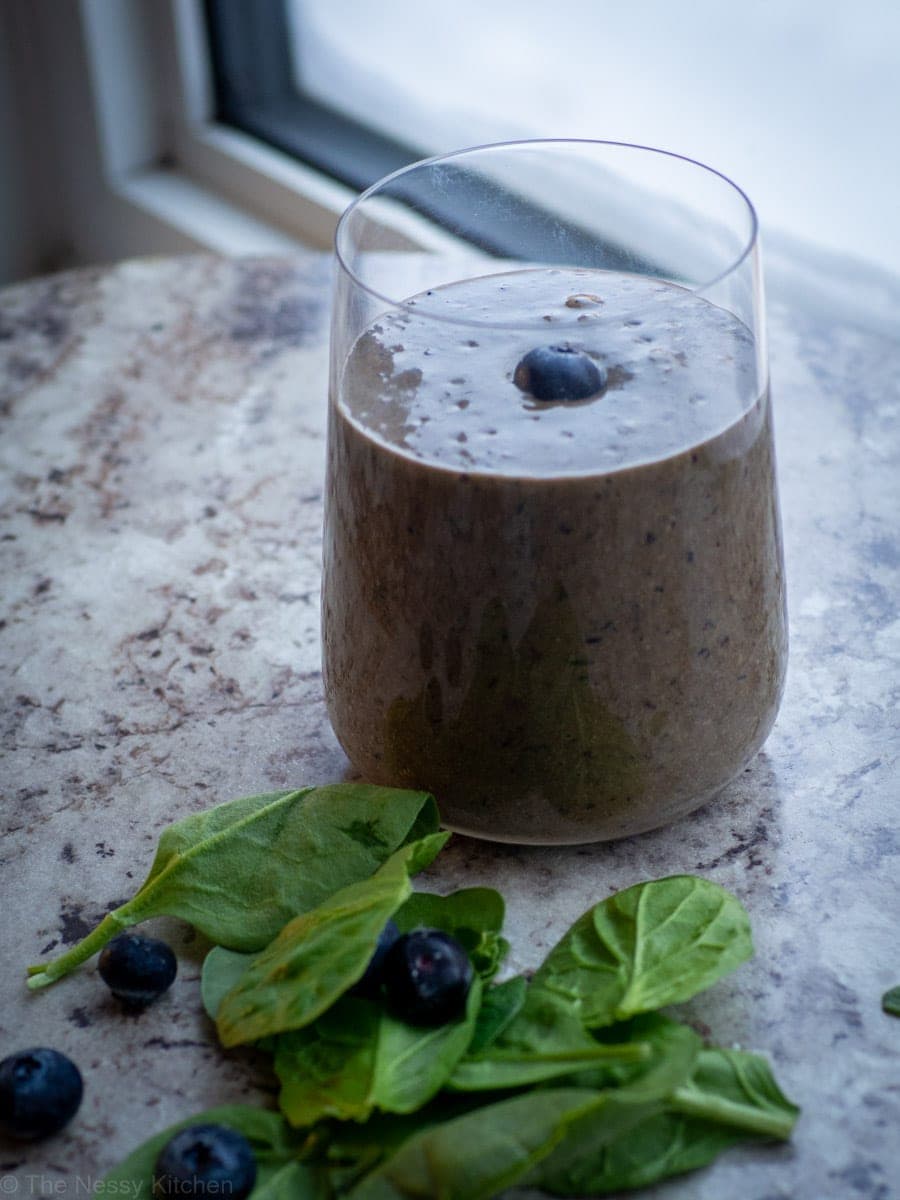 Glass with blueberry smoothie with spinach and blueberries on the counter next to it.