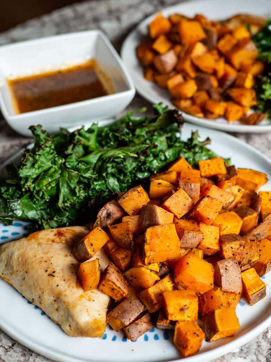 Plate with chicken breast, sweet potatoes and kale with sauce in a bowl in the background.