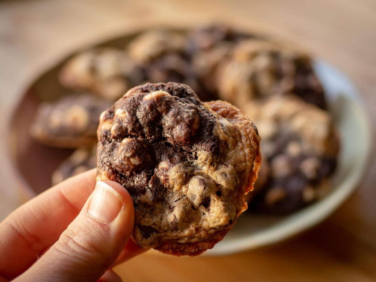 Hand holding a half chocolate chip. half chocolate white chocolate chip cookie with a plate of cookies in the background.