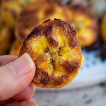 A baked ripe plantain round being held up with a plate of more in the background.
