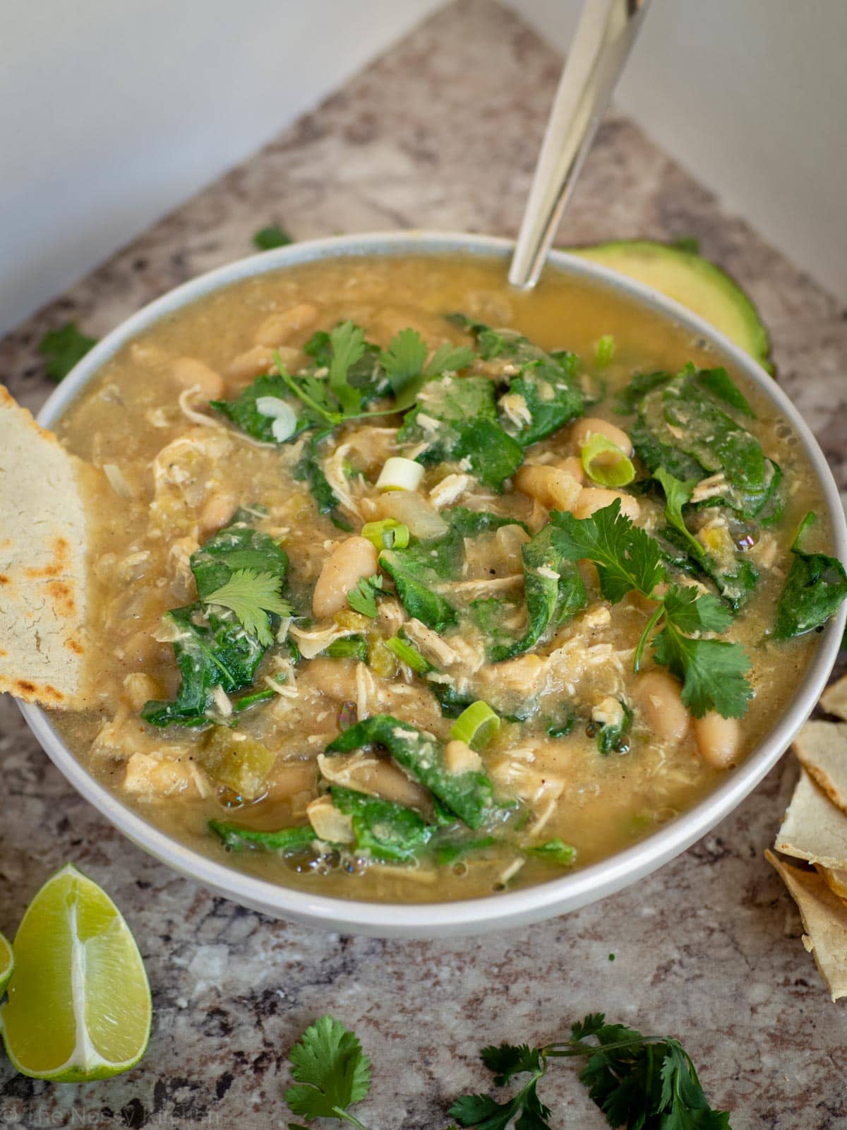 Bowl of white chicken chili with kale. Topped with cilantro and green onions with avocado and tortilla on the side.