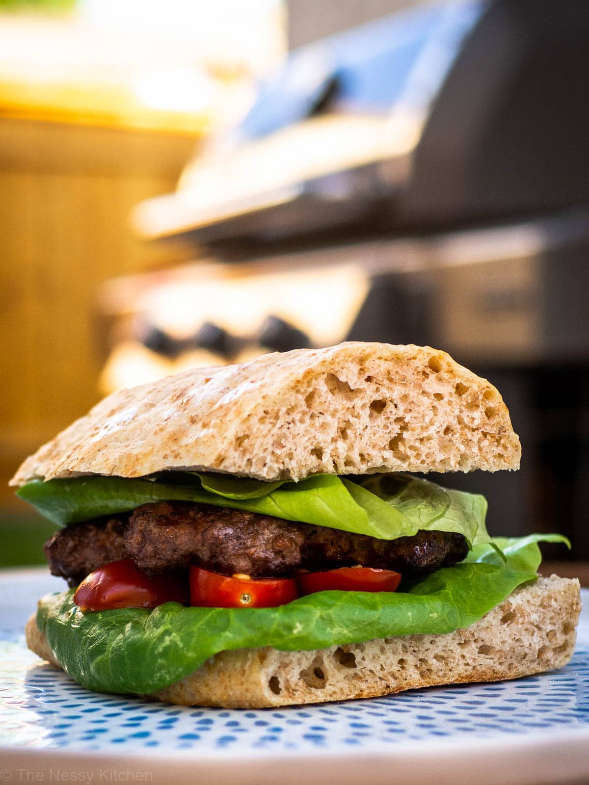 Barbecued beef burger on a bun with a barbecue grill in the background.