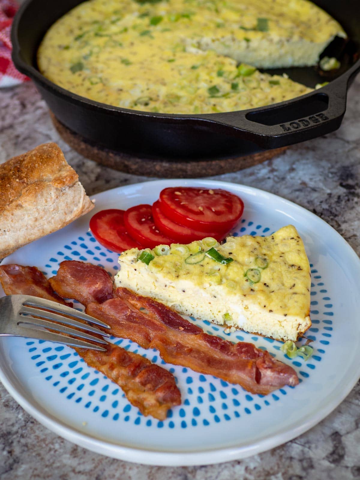 Frittata on a plate with crusty bread, sliced tomato and two strips of bacon.