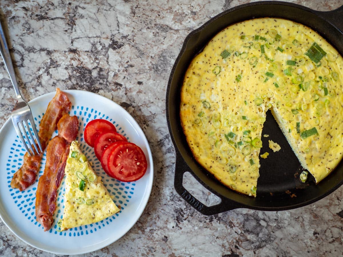 Slice of frittata on a plate with bacon and tomatoes next to a skillet with remaining frittata.