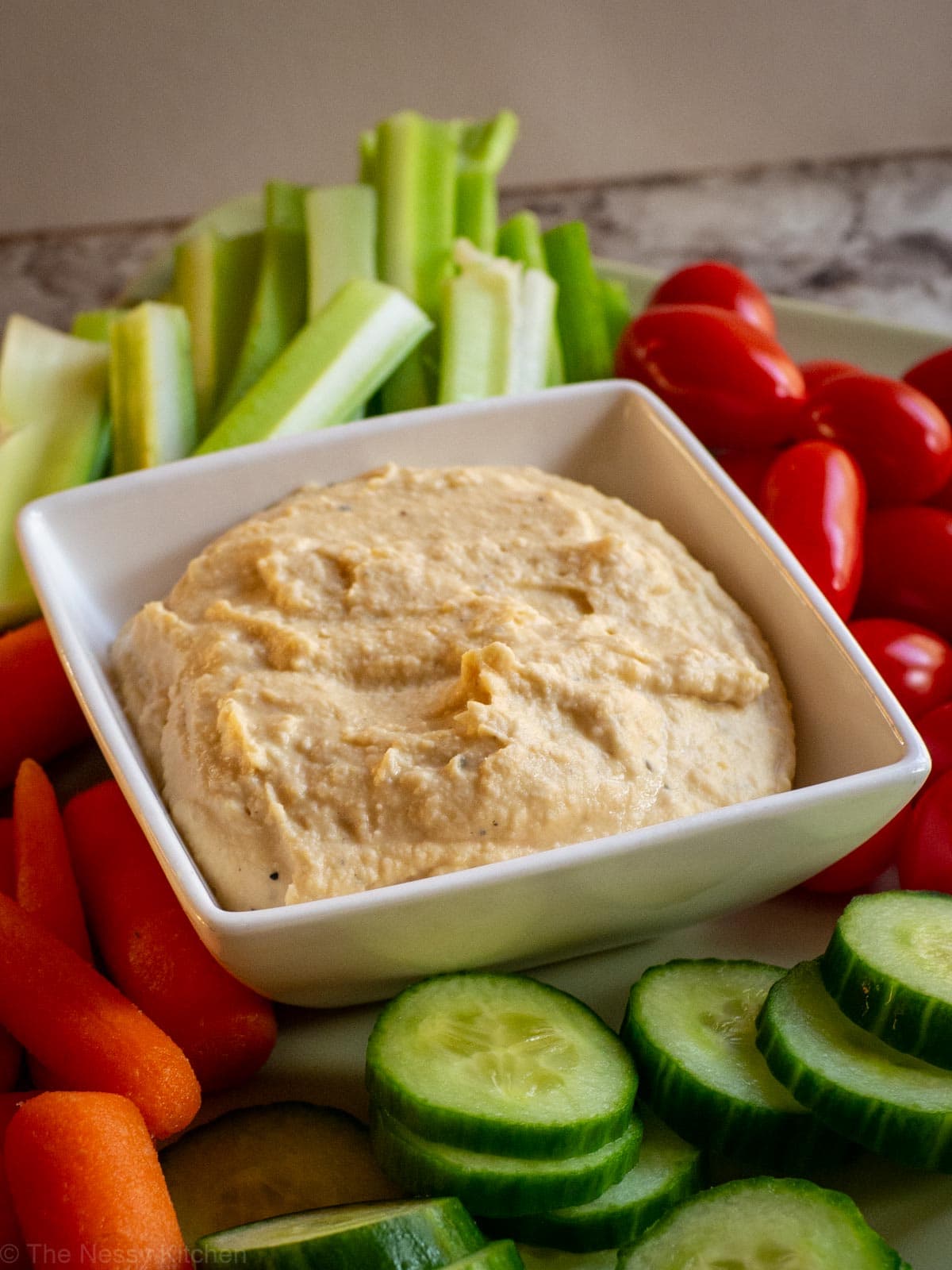 No garlic hummus in a white square bowl surrounded by celery, carrots, tomatoes and cucumbers.