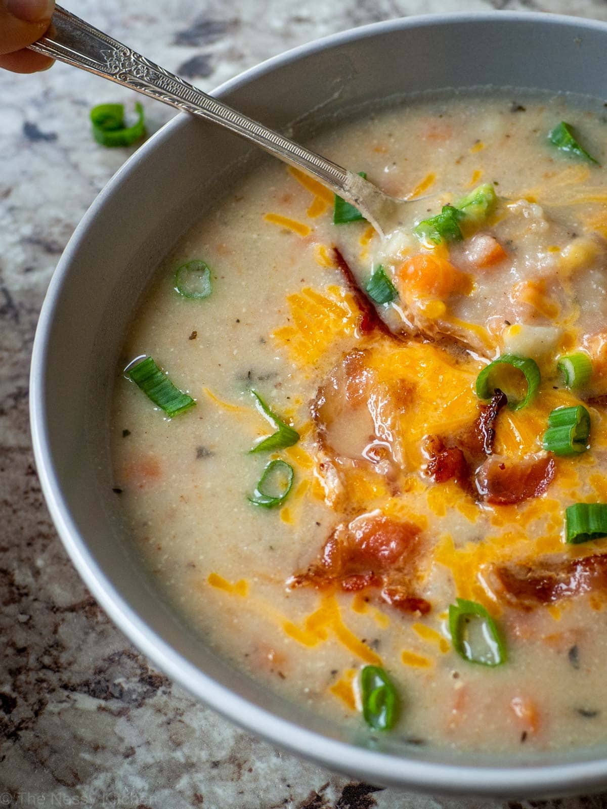 Spoon scooping a bite of potato soup from a grey bowl topped with green onions, cheddar cheese and bacon.