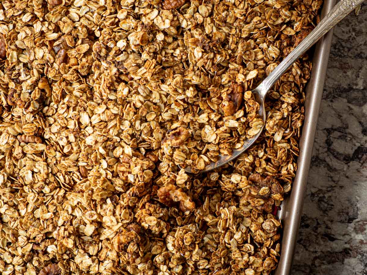 Cooked and cooled granola on a sheet pan being scooped with a large metal spoon.