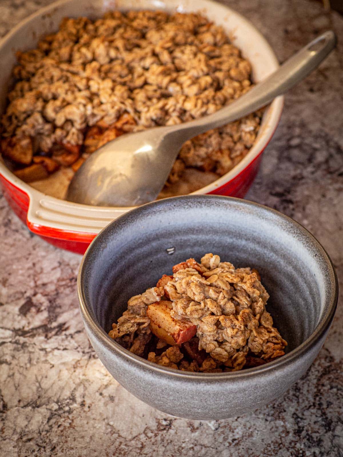 Apple crisp being scooped into a grey bowl from a red baking dish.