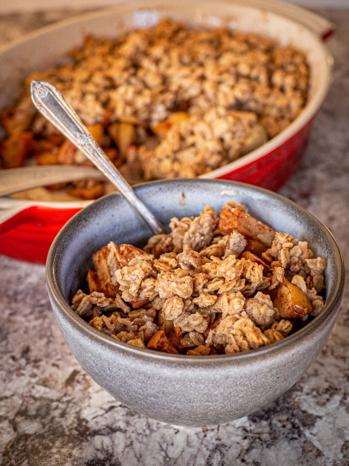 Bowl of apple crisp with a baking dish in the background.