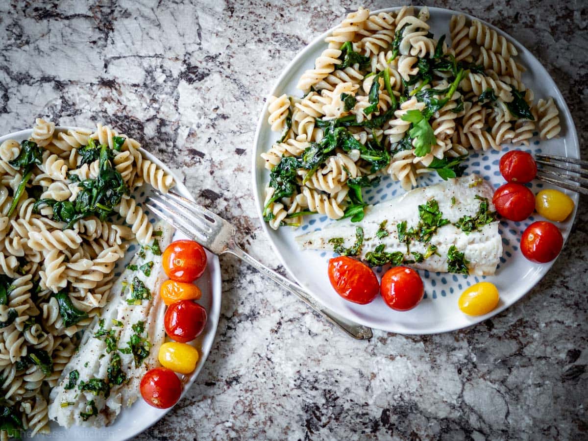 Top view of two plates of white fish with tomatoes alongside pasta.