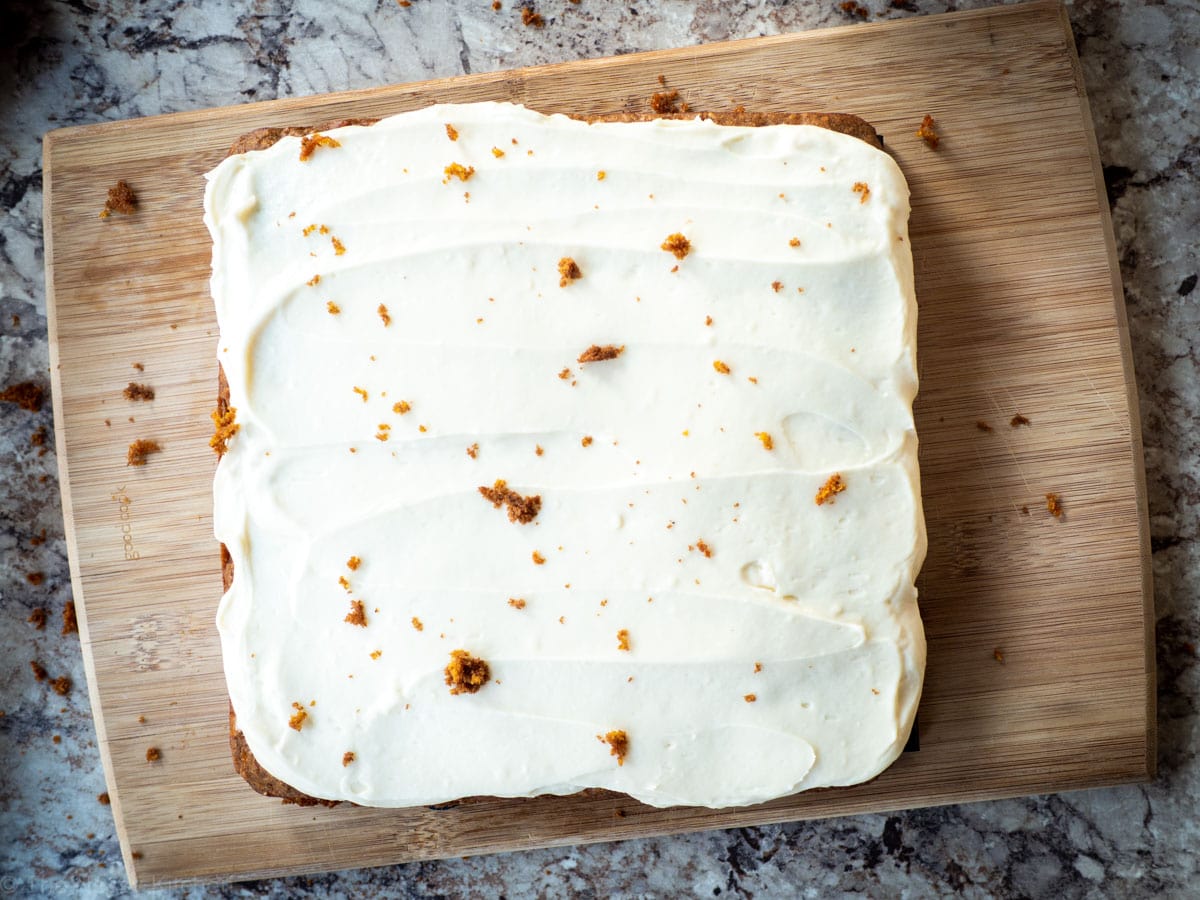 Frosted low sugar carrot cake on a wooden cutting board and sprinkled with cake crumbs.