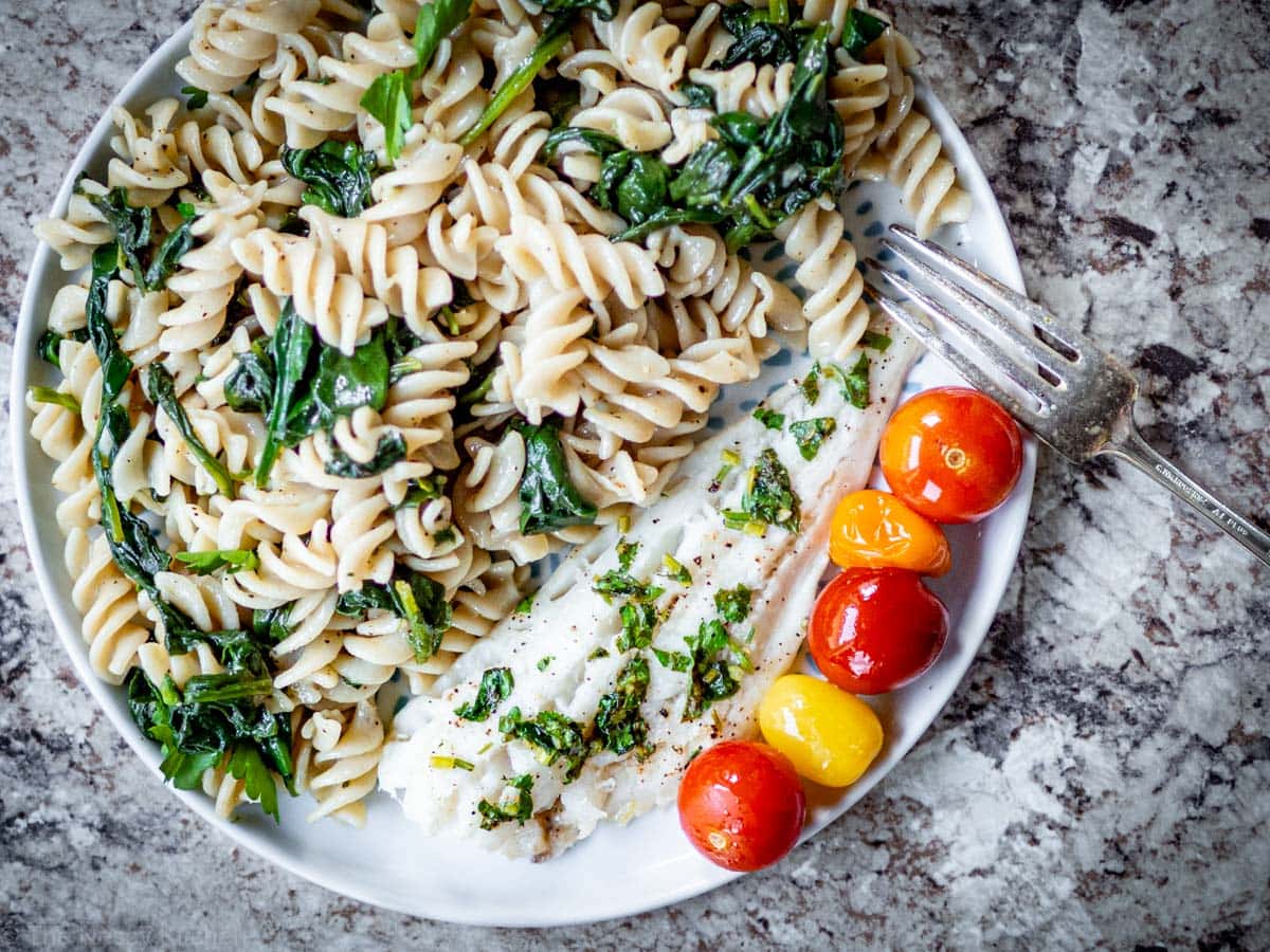 Top view of a plate with pasta, roasted white fish and tomatoes with a fork on the side.