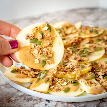 Hand holding up an apple slice topped with nut butter and granola.