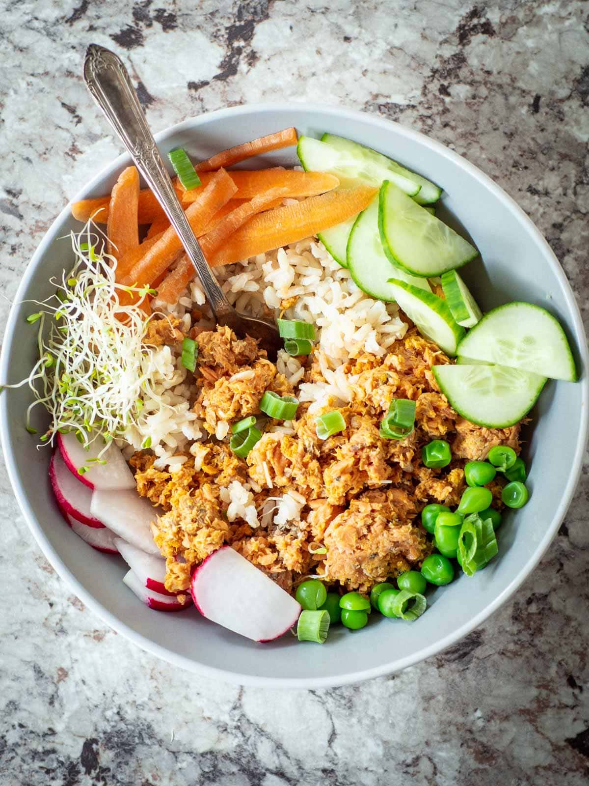 Top view of a bowl of canned salmon over rice topped with cucumbers, radishes, carrots, alfalfa and green onions.