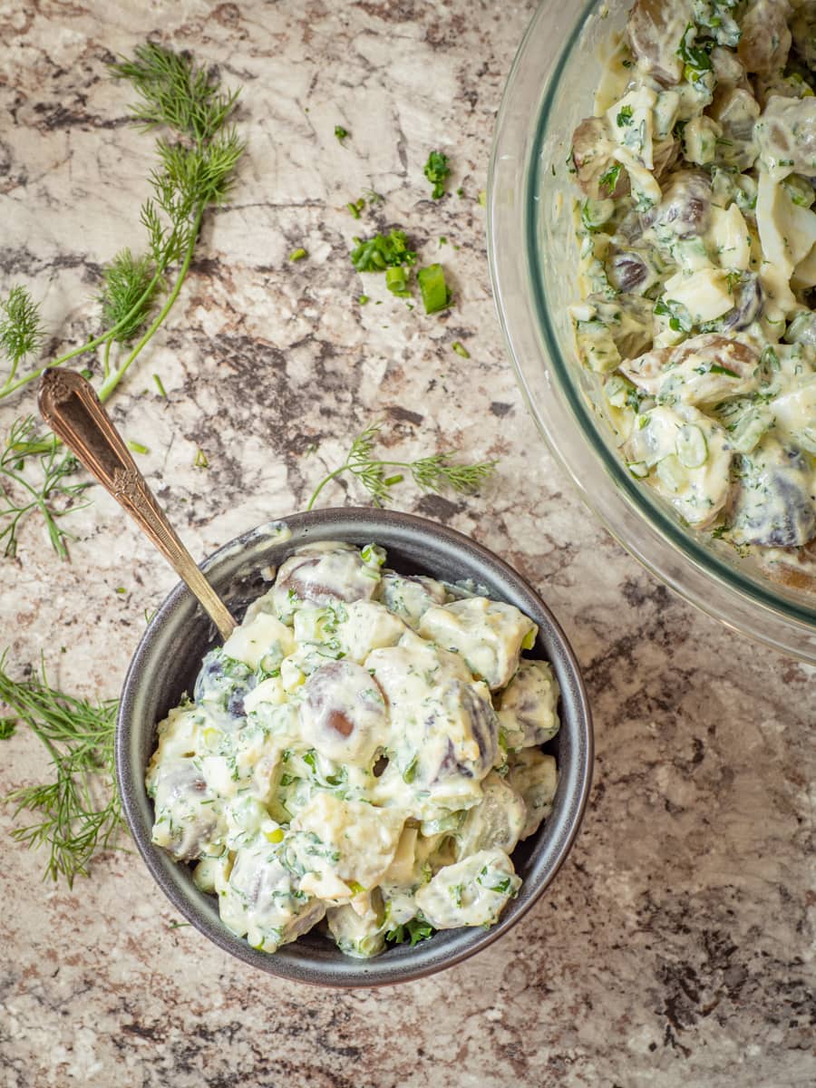 Small bowl of dished out potato salad in a grey bowl with fresh herbs on the countertop.