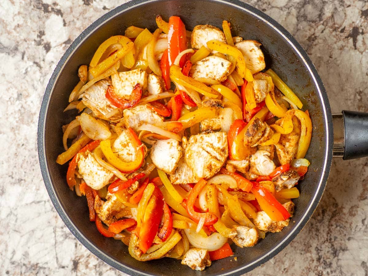 Chicken stirred into a frying pan with peppers and onions.