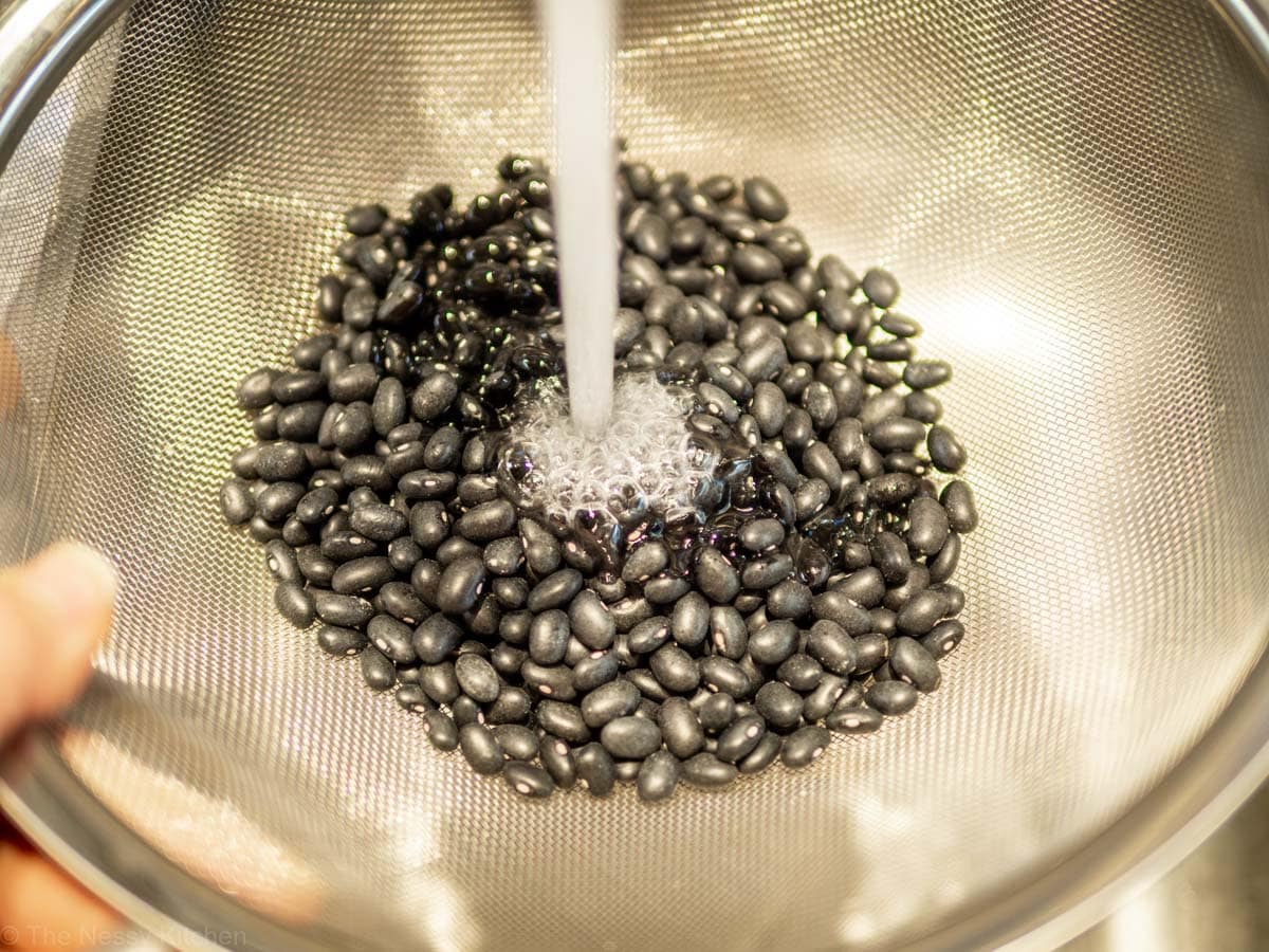 Dry black beans being rinsed in a sieve.