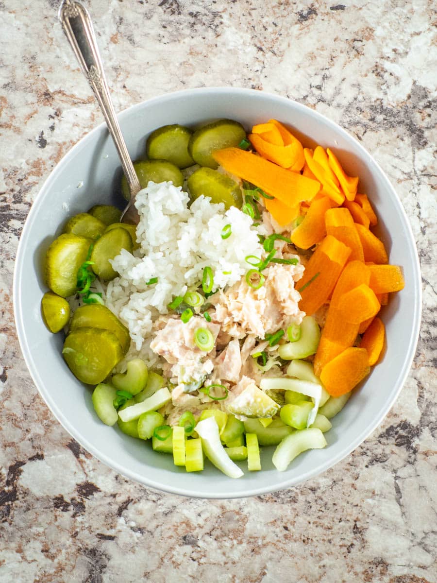 Tuna and rice in a bowl topped with pickles and fresh vegetables.