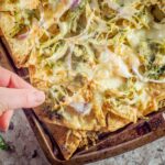 Hand taking a tortilla chip from a tray of chimichurri chicken nachos.